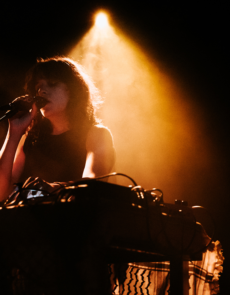 Snakeskin's vocalist Julia Sabra on stage, singing in concentration, while a spotlight shines just behind her.